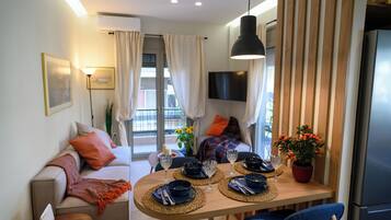 A modern living room with a sofa, dining table, and chairs. There is a TV mounted on the wall, a refrigerator, and a window with curtains.
