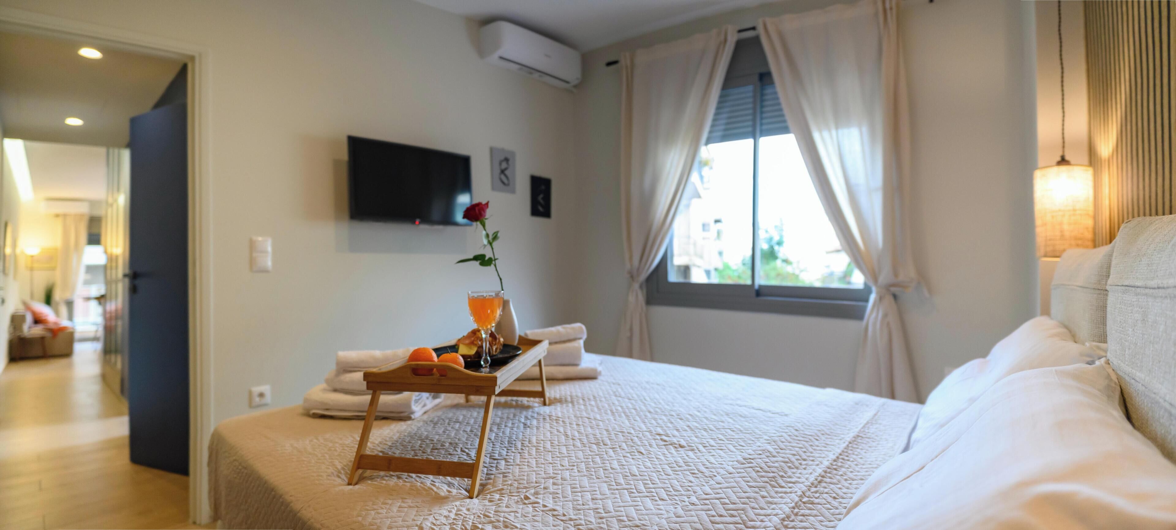 Habitación de hotel con una cama, una mesita de noche con una bandeja de frutas, un florero con una sola flor, un televisor de pantalla plana montado en la pared y una ventana con cortinas.