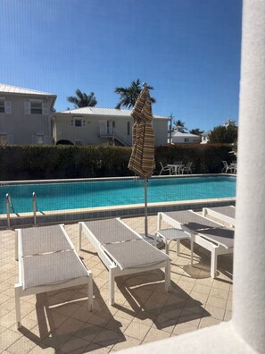 Un área junto a la piscina con sillones de descanso y una sombrilla, una piscina azul cristalina y un edificio al fondo.