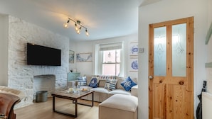 A living room with a stone wall, a sofa with nautical-themed cushions, a wooden coffee table, and a television mounted on the wall. - 3 Bed in Brixham (oc-p30777) (Brixham)