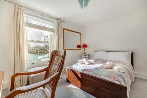 A bedroom with a wooden bed, a chair, a bedside table with a lamp, and a window with curtains.