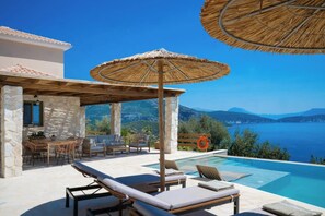 Un espace aménagé au bord de la piscine, avec des chaises longues, une table et des parasols, offrant une vue sur un plan d’eau et des montagnes.