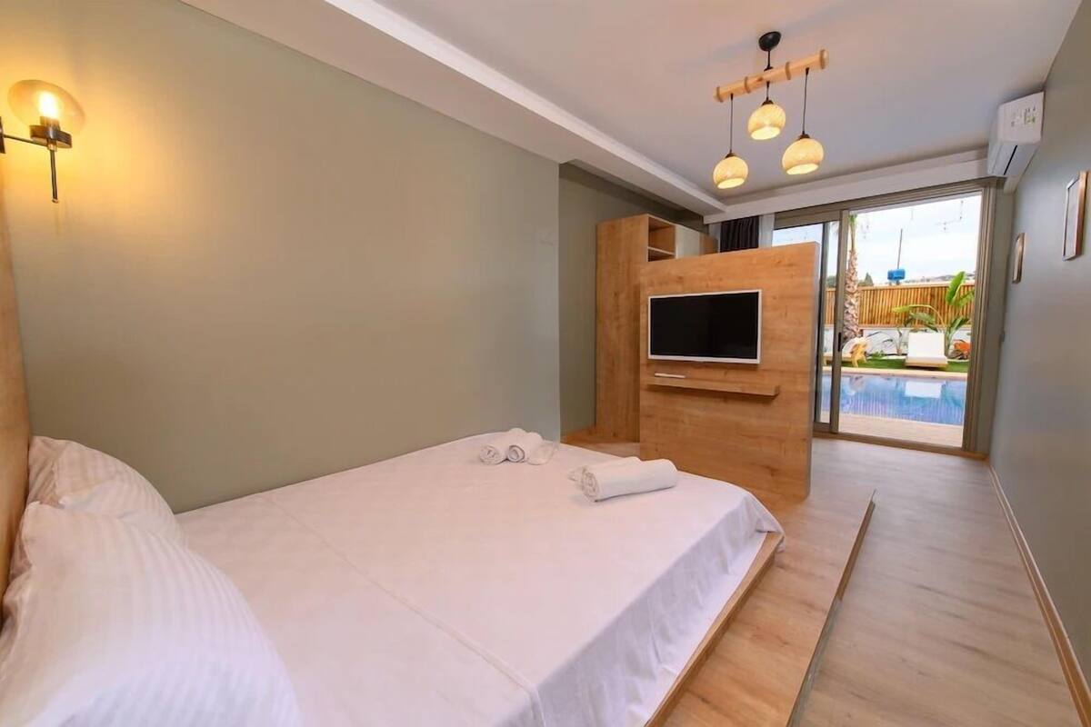 A hotel room with a bed, a TV, a wooden cabinet, and a view of an outdoor swimming pool.
