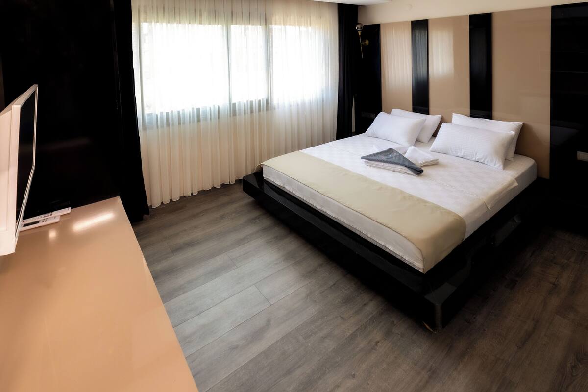 A modern hotel room with a large bed, a flat-screen TV, and a window with sheer curtains.