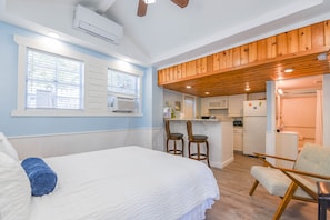 Quarto com cama, duas cadeiras, cozinha compacta e teto de madeira.