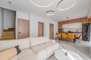 A modern living room with a sofa, a dining area with yellow chairs, a kitchen with wooden cabinets, and a staircase.