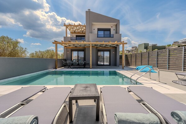 Et moderne poolområde med liggestole, et pergola og en toetagers bygning med store vinduer.