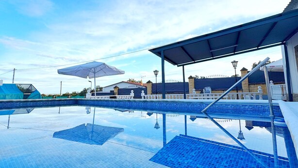 A swimming pool with clear blue tiles, a covered seating area, and a view of buildings in the background. - La Colina Rural House El Duque Accommodations (Córdoba)