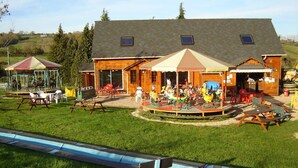 A wooden building with a covered patio area, picnic tables, and a carousel. - Rental of comfortable trailers for 4 people at DEVALPARC leisure park (Livarot-Pays-d'Auge)