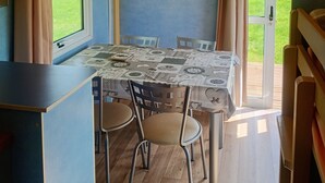 A cozy dining area with a table set for four, a wooden ceiling, and a view of a green landscape through a glass door. - Rental of comfortable trailers for 4 people at DEVALPARC leisure park (Livarot-Pays-d'Auge)