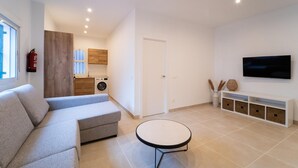Ein modernes Wohnzimmer mit grauem Sofa, rundem Couchtisch, Wand-Fernseher und Küchenzeile inklusive Waschmaschine.