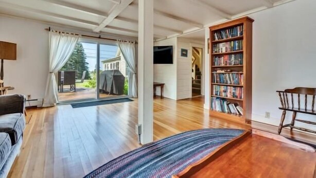 A spacious room with a wooden floor, a bookshelf, a sofa, and a large window with curtains. - Seaside Farmhouse | Cozy Waterfront Getaway in Ten (Saint George)
