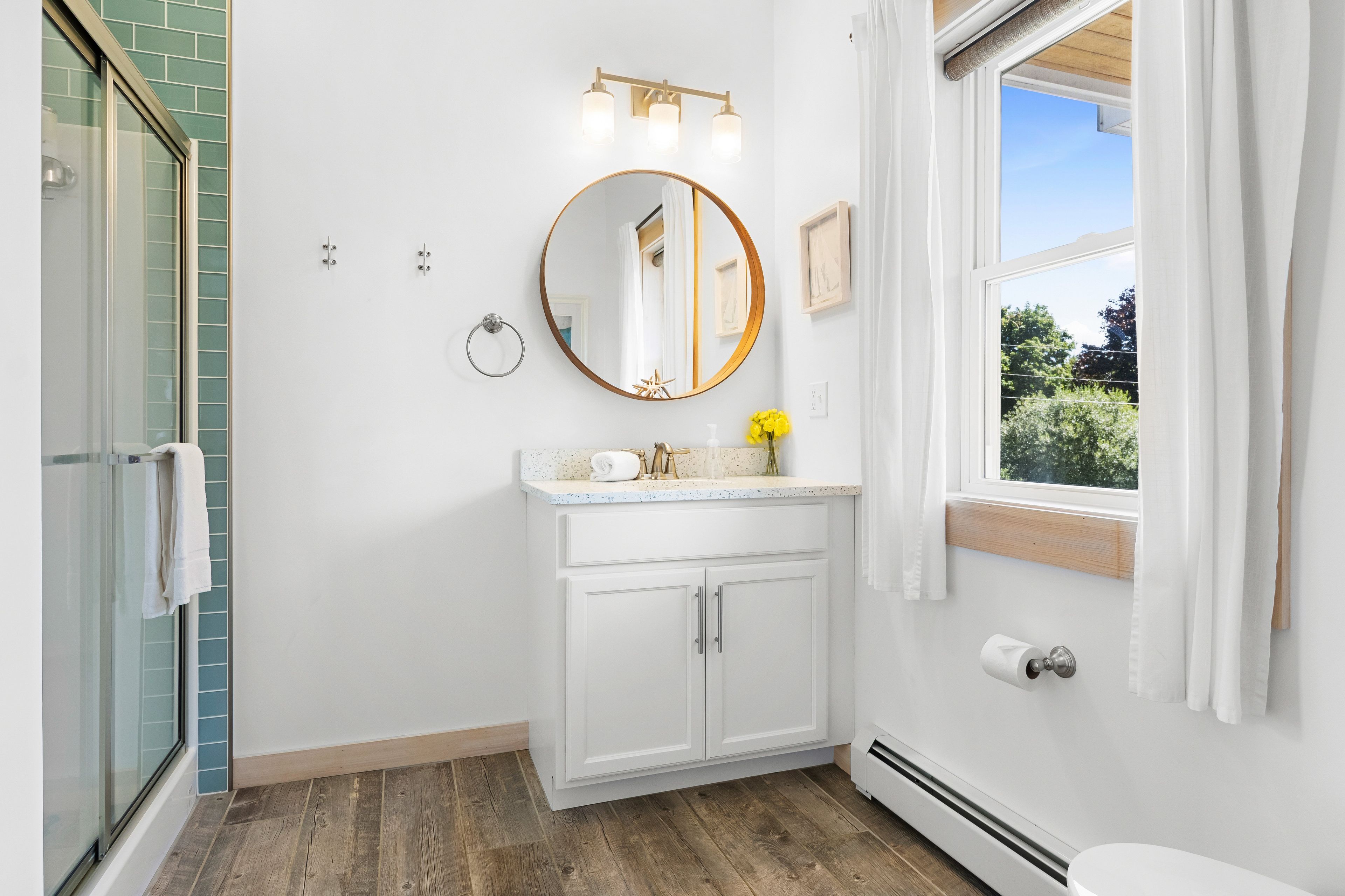 Une salle de bain dotée d’une douche à parois de verre, d’un meuble-lavabo blanc avec un miroir rond et d’une fenêtre donnant sur un paysage verdoyant.