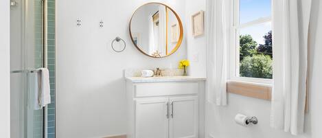 Une salle de bain dotée d’une douche à parois de verre, d’un meuble-lavabo blanc avec un miroir rond et d’une fenêtre donnant sur un paysage verdoyant.