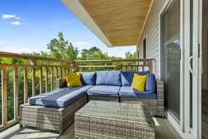 A covered outdoor seating area with a sofa and ottomans, a wooden deck, and a view of trees.