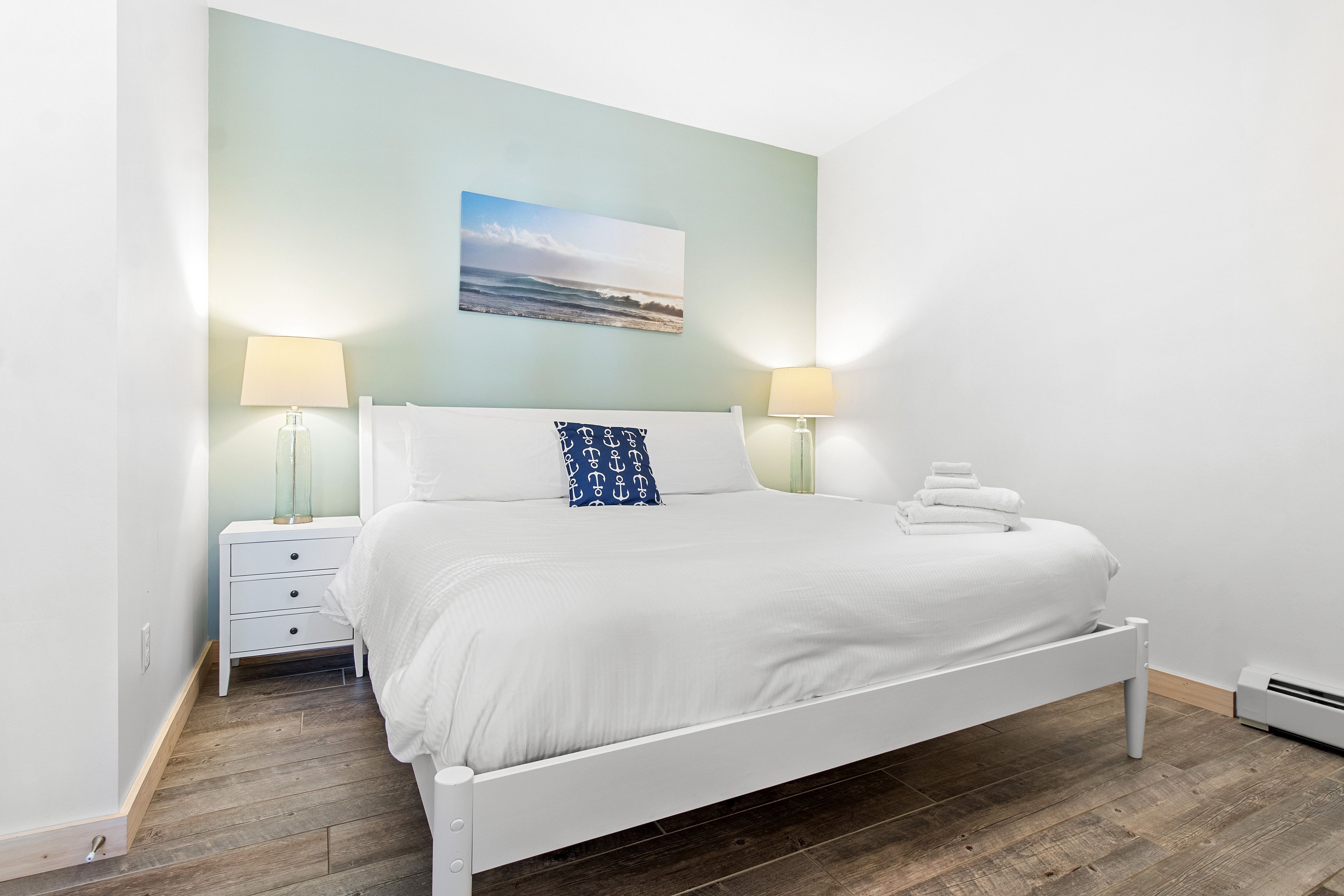 A neatly made bed with white bedding and a single blue pillow with anchor patterns, flanked by two bedside tables with lamps, in a room with a wooden floor and a light blue wall.