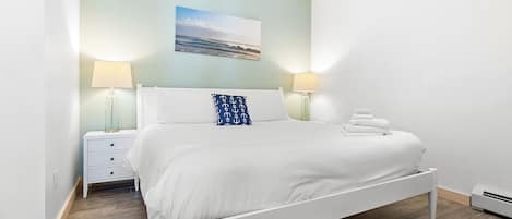 A neatly made bed with white bedding and a single blue pillow with anchor patterns, flanked by two bedside tables with lamps, in a room with a wooden floor and a light blue wall.