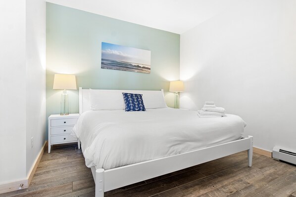 A neatly made bed with white bedding and a single blue pillow with anchor patterns, flanked by two bedside tables with lamps, in a room with a wooden floor and a light blue wall.