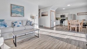 A modern living room with a sofa, coffee table, and artwork. A kitchen with a dining area is visible in the background. - Ocean Breeze Haven - Short walk from Crescent Beac (Owls Head)