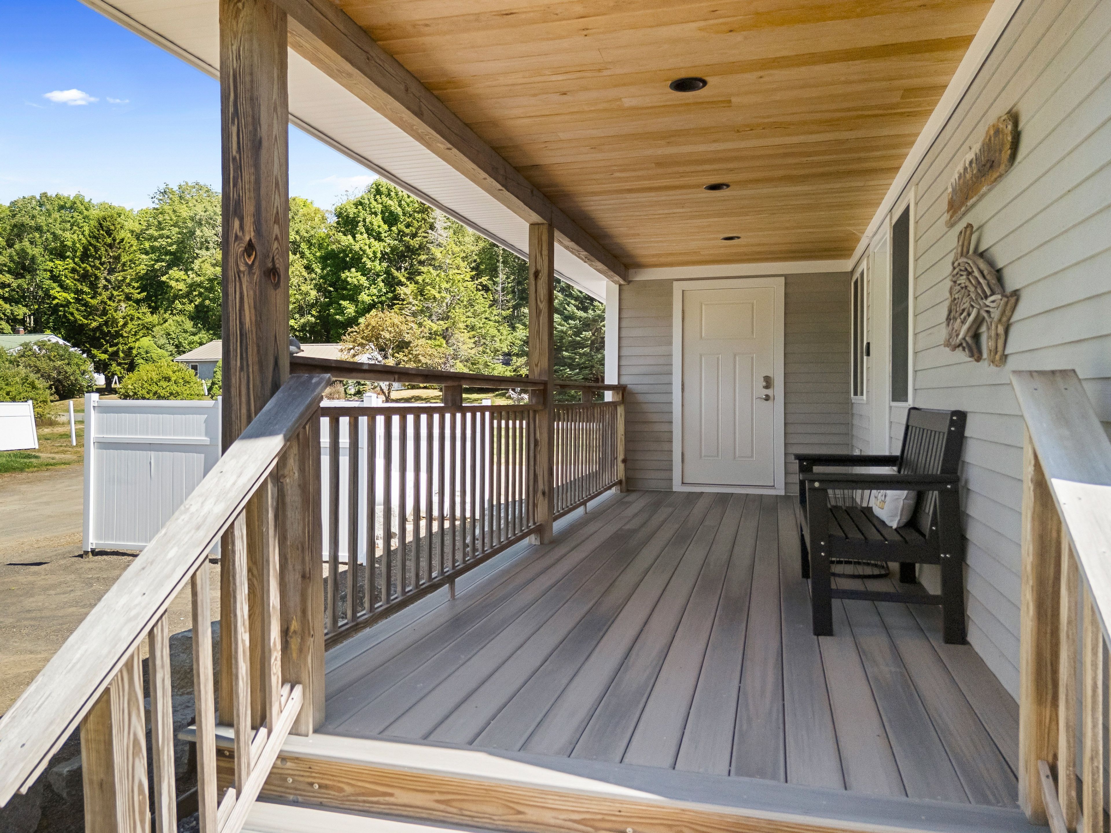 Une terrasse en bois avec un banc et une porte, entourée d’une clôture blanche et d’arbres.