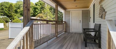 Une terrasse en bois avec un banc et une porte, entourée d’une clôture blanche et d’arbres.