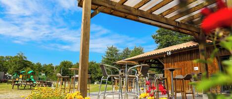 A wooden deck with metal chairs and tables, a covered seating area, and a playground in the background.
