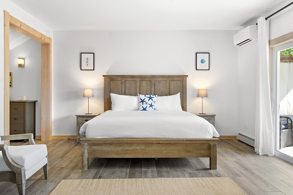 A bedroom with a wooden bed, two bedside lamps, a nightstand, a chair, and a large mirror. - Ocean Breeze East Wind – Modern 2BR/2BA home by Cr (Owls Head)