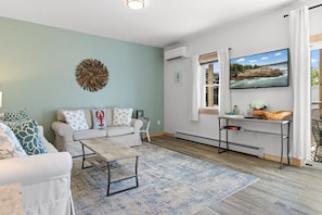 A modern living room with a sofa set, a coffee table, a flat-screen TV, and a view of the outdoors through a window. - Ocean Breeze East Wind – Modern 2BR/2BA home by Cr (Owls Head)