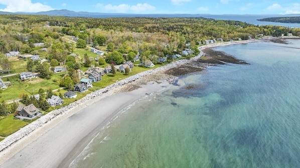 Vue aérienne d’une zone résidentielle côtière, avec des maisons le long d’une plage, une eau claire et un paysage boisé en arrière-plan.