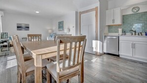 Un coin repas avec une table et des chaises en bois, une cuisine avec des meubles blancs et un plan de travail vert, et une horloge au mur.