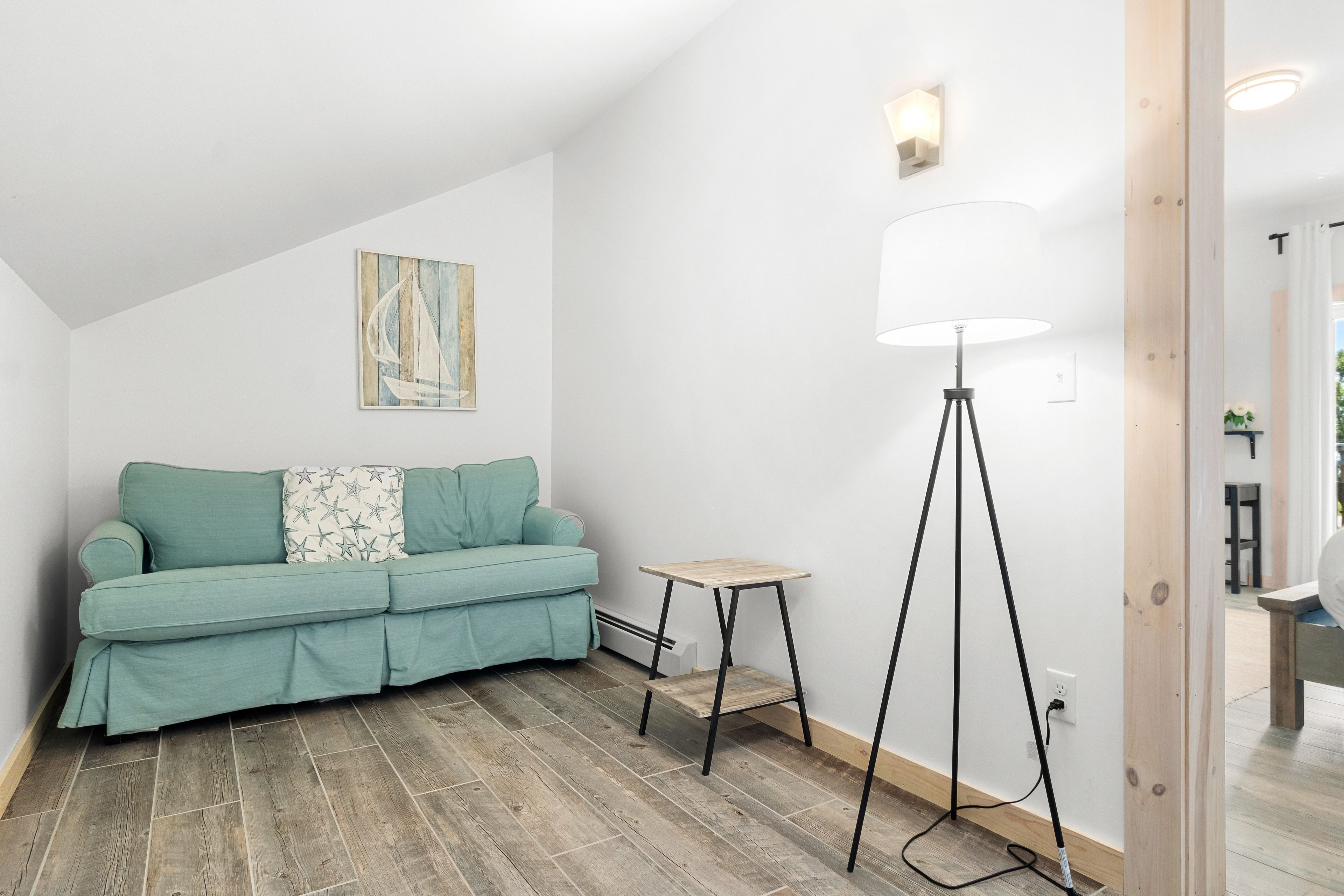 A modern living room with a teal sofa, a small wooden side table, a floor lamp, and a painting on the wall.