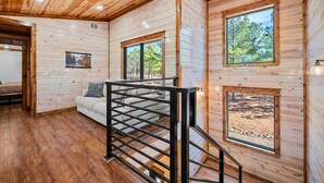 Interior de una cabaña de madera con una escalera, un sofá blanco y ventanas amplias que ofrecen vistas a árboles.