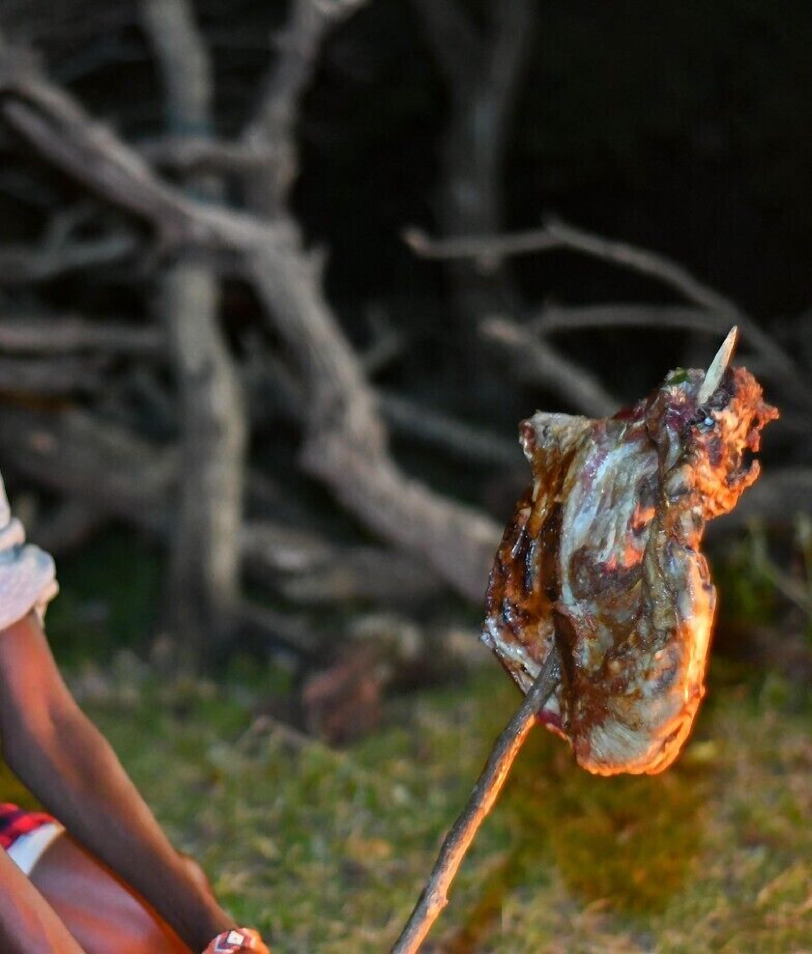 A person holding a skewered meat, likely being grilled, outdoors with bare trees in the background.