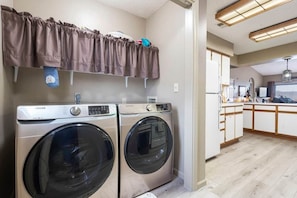 A laundry area with two front-loading washing machines, a hanging rack, and a kitchen area with wooden cabinets and a countertop.