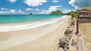A beach with clear turquoise waters, a sandy shore, and a stone walkway leading to a covered seating area with umbrellas. - La Sirène du Diamant  (Le Diamant)