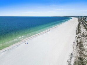 An aerial view of a beach with a long stretch of white sand, clear blue water, and a row of buildings along the shore. - 3BR Retreat: Pool, Patio & Steps from Beach (Mexico Beach)