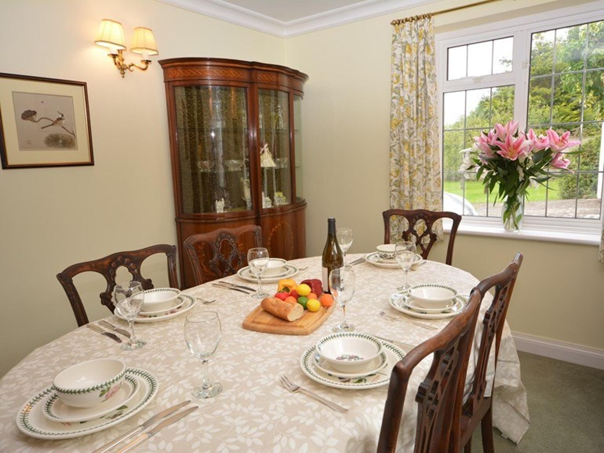 A dining room with a round table set for four, surrounded by wooden chairs. A wooden sideboard with glass doors is against the wall, and a window with floral curtains is visible.