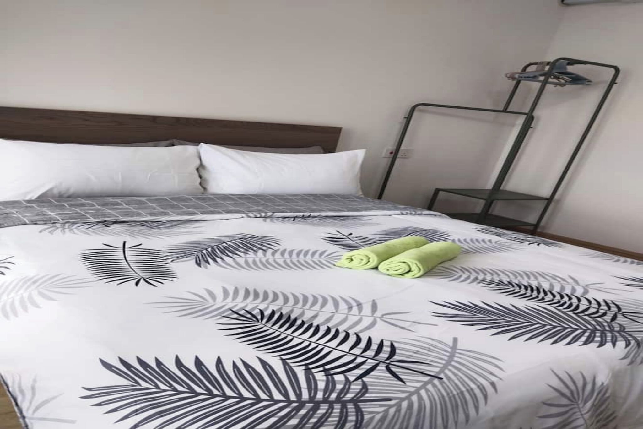 Ein ordentlich bezogenes Bett mit einem gemusterten Überwurf, zwei aufgerollte Handtücher und ein Metallregal an der Wand.