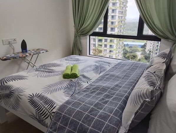 Una cama bien hecha con una colcha estampada con motivos de hojas, un par de toallas enrolladas y una ventana con cortinas.