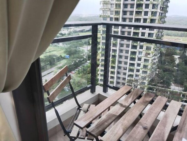 Un balcón con un banco de madera y una silla de metal, con vistas a un paisaje urbano con rascacielos y zonas verdes.