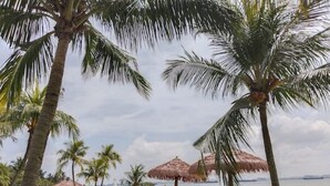 A beach with palm trees, a thatched-roof structure, and a sandy area with lounge chairs. - Holiday Home@Forest City~TV Box~2BR1B 4-6pax 1807 (Gelang Patah)
