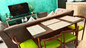 Un área de comedor con una mesa de madera y sillas con cojines verdes, un sofá y un televisor sobre un mueble.