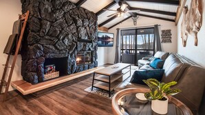 A cozy living room with a stone fireplace, a wooden bench, a leather sofa, and a large window offering a view of trees. - Breathtaking Lake Views | Private Dock, Fire Pit & Chef’s Kitchen (Hauser)