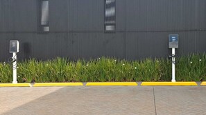 Two electric vehicle charging stations in front of a building with a corrugated metal facade and a row of green plants. - JD Hotel cravinhos (Cravinhos)