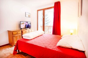 Habitació d'hotel amb un llit, una petita arca de fusta, una televisió i un balcó amb cortines vermelles.