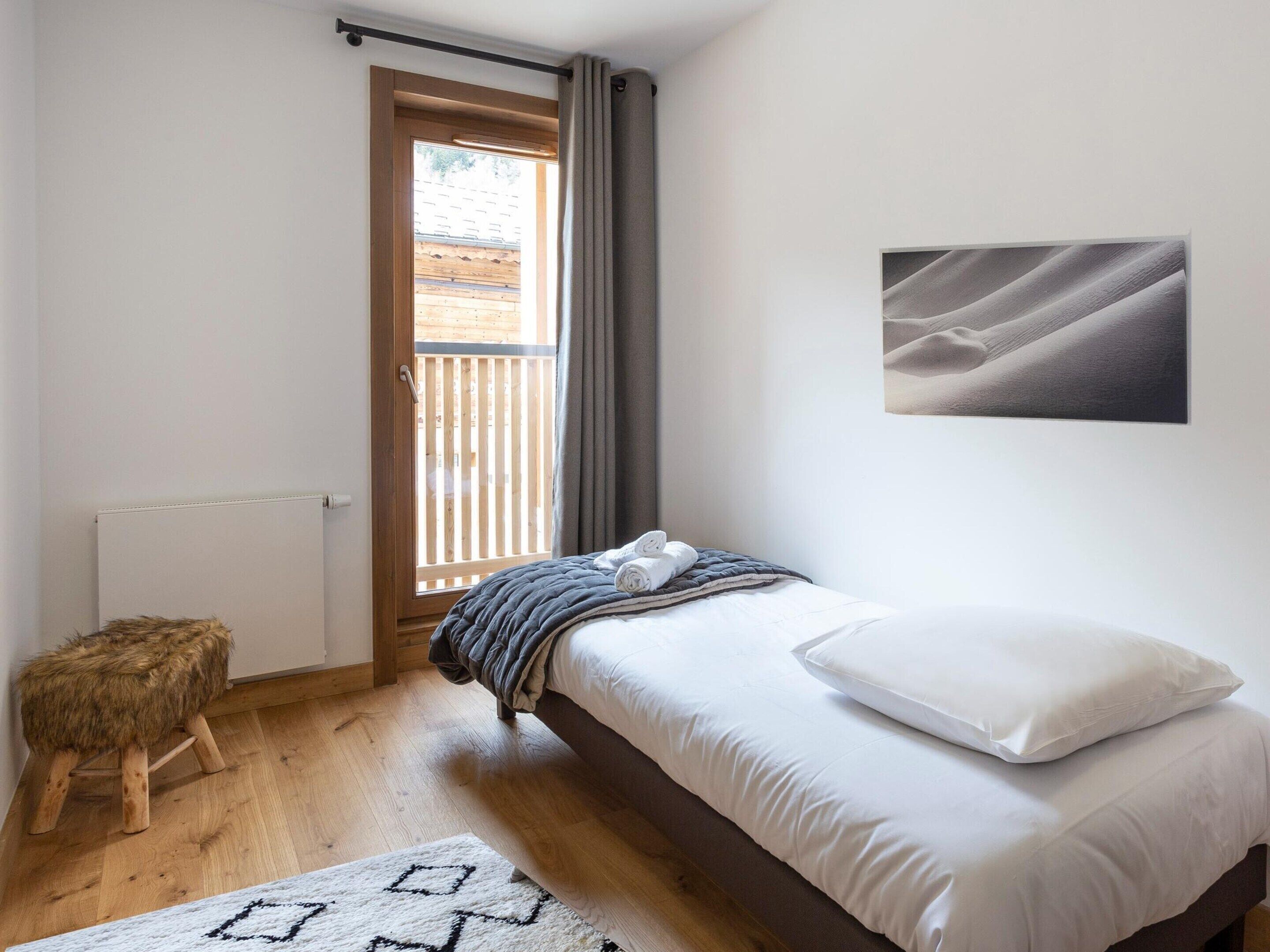 Une chambre à coucher avec un lit, un tabouret en bois, un tapis blanc, une fenêtre avec des rideaux et un tableau accroché au mur.