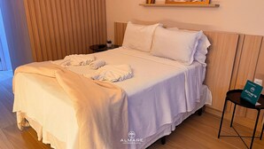Una cama bien hecha con edredón blanco y almohadas, una mesita de noche con una planta y un cartel que indica ALMARE HOTEL & FLATS.