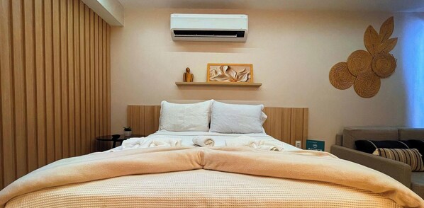 A hotel room with a bed, an air conditioner, a bedside table, a wall-mounted shelf with decorative items, and a wall art piece.