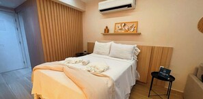 En pænt redt seng med et beige tæppe, et lille natbord med en bog, og en vægmonteret aircondition.