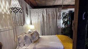A bedroom with a bed, white curtains, a hanging macrame light fixture, and a bedside lamp. - The Sunburst Oasis  (Palm Harbor)